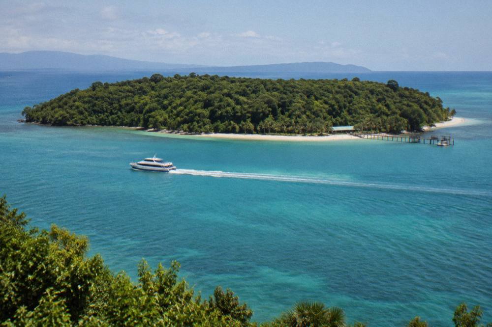 A fast catamaran ferry crossing calm turquoise water toward a green coral island
